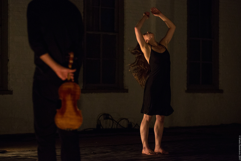 A woman in a black tank dress raises her arms above her head. A man dressed all in black holds a violin behind her back.